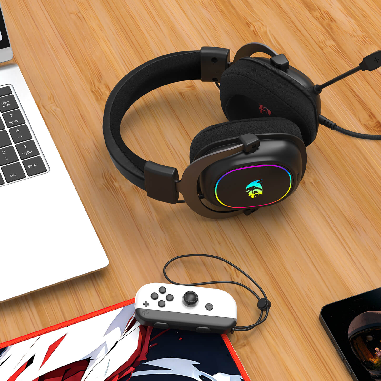 Gaming headset with RGB lighting on a wooden desk next to a laptop and gaming console.