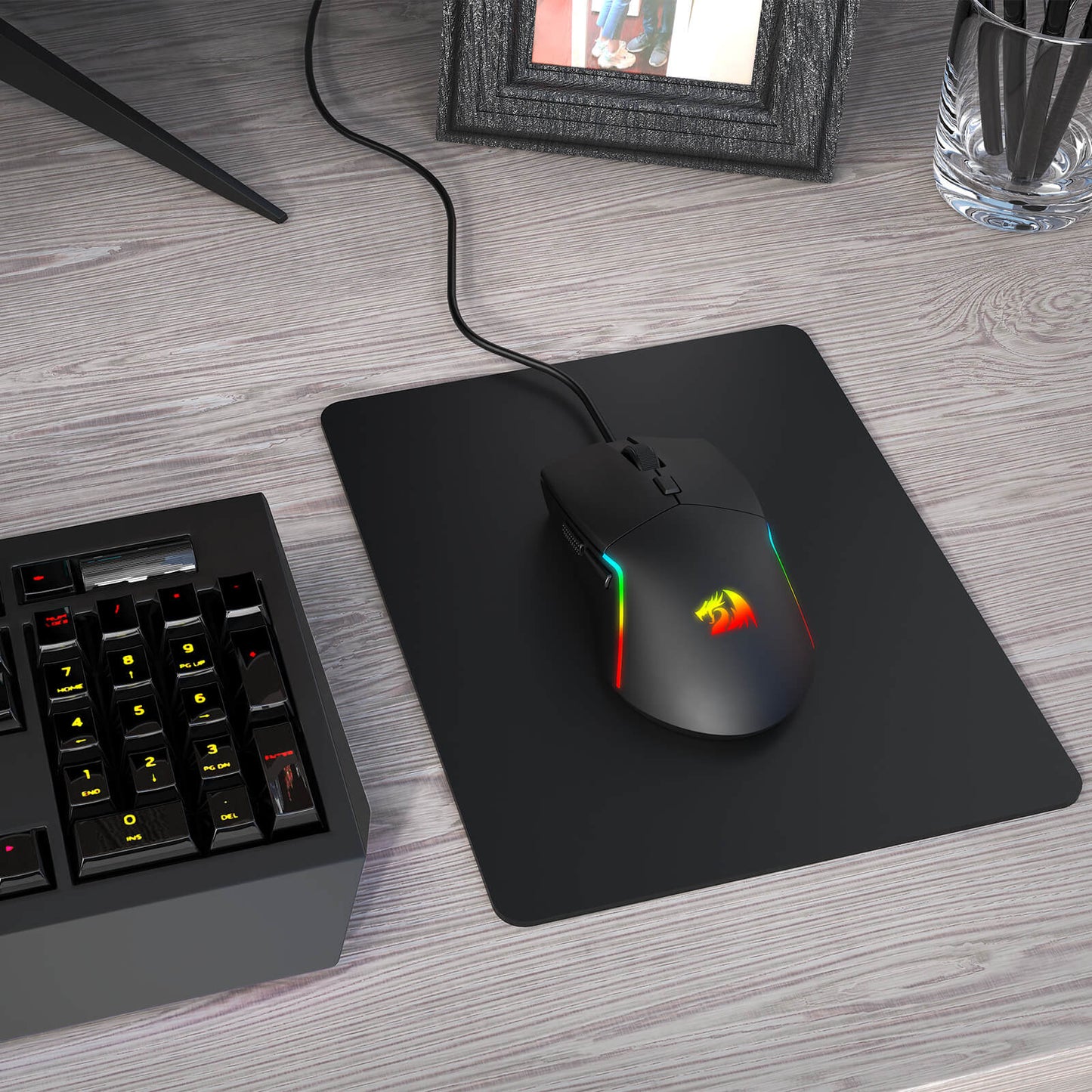 A Redragon RGB gaming mouse with a glowing logo and side strips rests on a black mouse pad next to a mechanical keyboard.