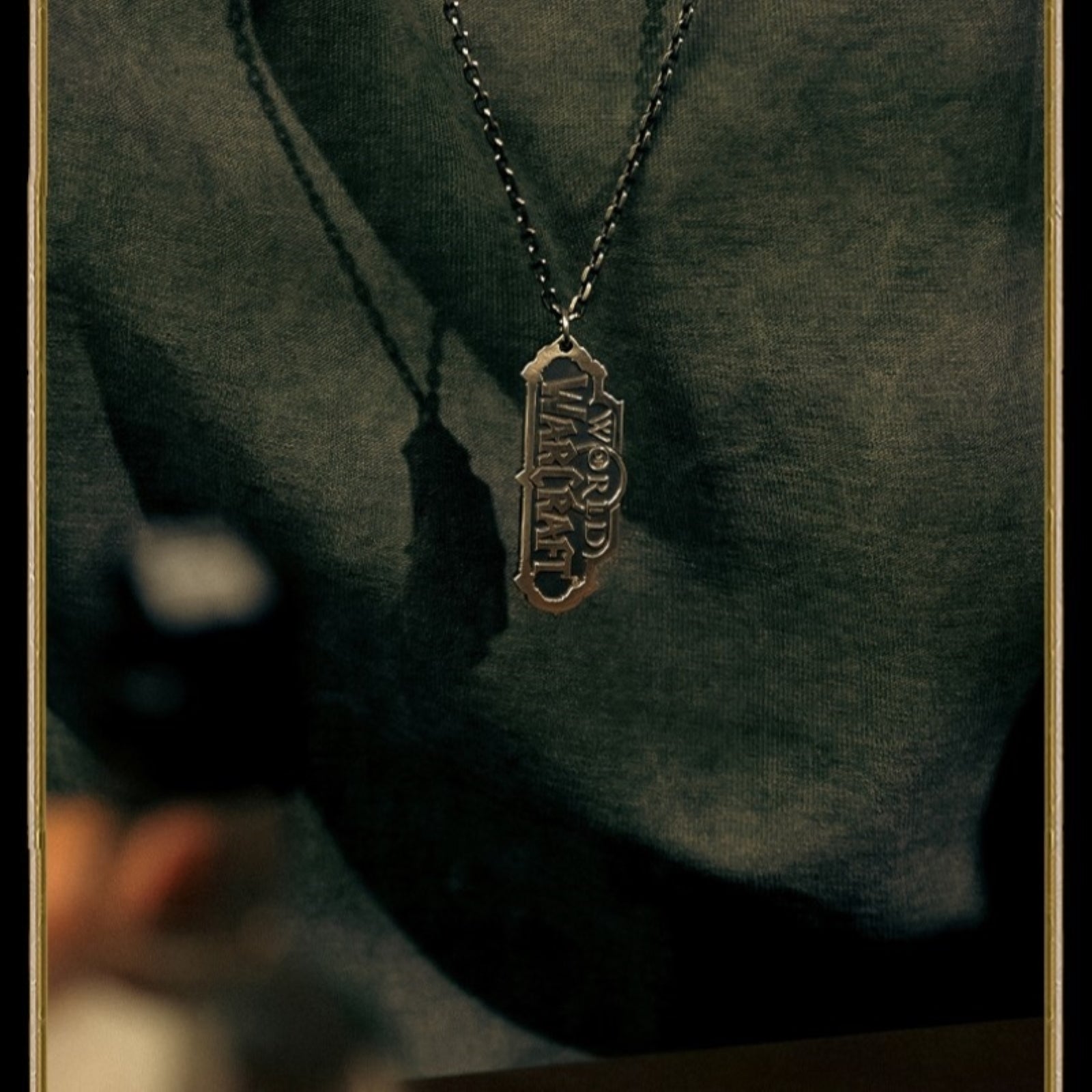 A metallic "WORLD OF WARCRAFT" pendant (in its iconic stylized font) hangs from a chain against the textured fabric of a dark green garment, casting a soft shadow.