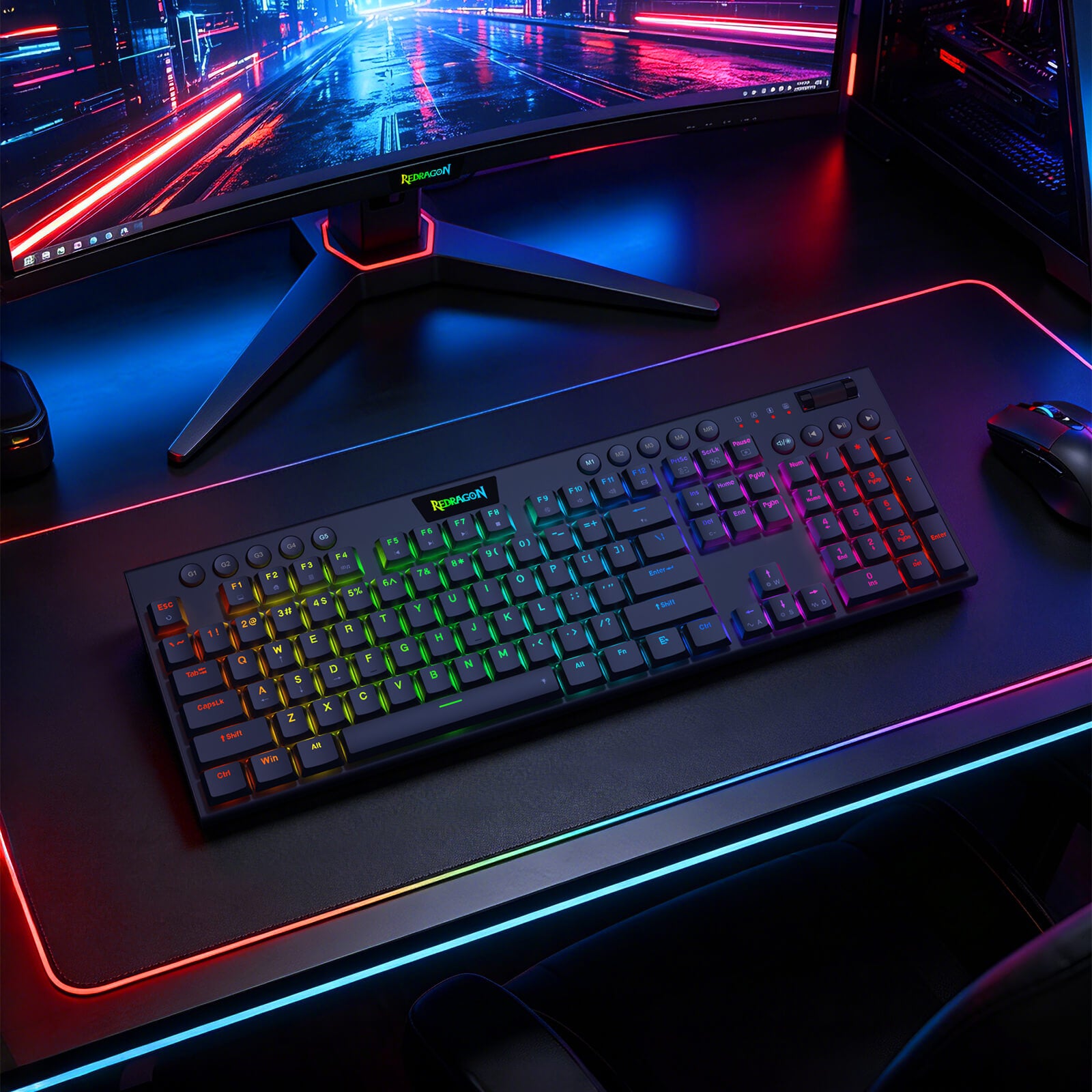 A black mechanical gaming keyboard with RGB backlighting sits on a matching black desk mat with a glowing multicolor LED border, set on a desk under a computer monitor and next to a mouse.