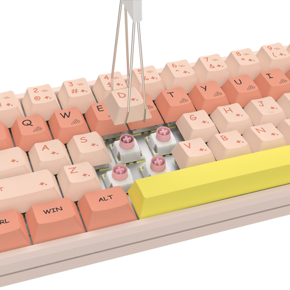 A close-up of a mechanical keyboard with pastel peach, cream, and coral keycaps, a large bright yellow spacebar, and visible pink switch capsules in the switch area. A metal key-stripping tool is positioned above the keyboard against a clean white background.