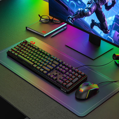 A Redragon RGB mechanical keyboard and gaming mouse are displayed on a branded desk mat in a modern gaming setup, with a monitor and webcam in the background.