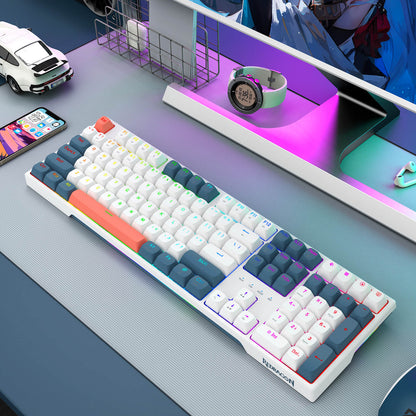 A white mechanical keyboard with colorful RGB backlighting and custom keycaps in shades of blue, teal, orange, and purple, shown on a gray desk next to a smartphone and a toy car.