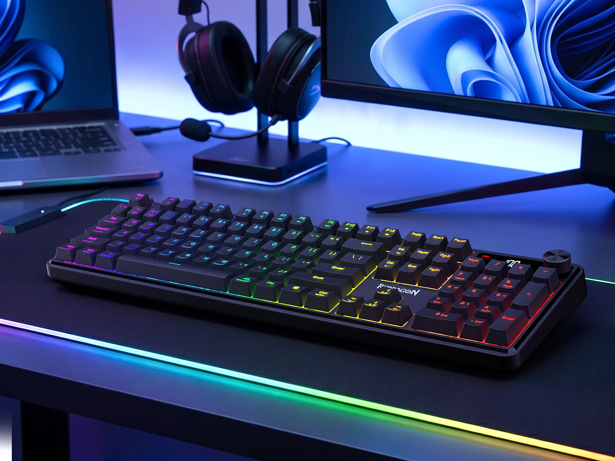 A black mechanical gaming keyboard with RGB backlighting sits on a desk in a styled setup, with a monitor, headphones on a stand, and a laptop visible in the background under blue and multicolor lighting.